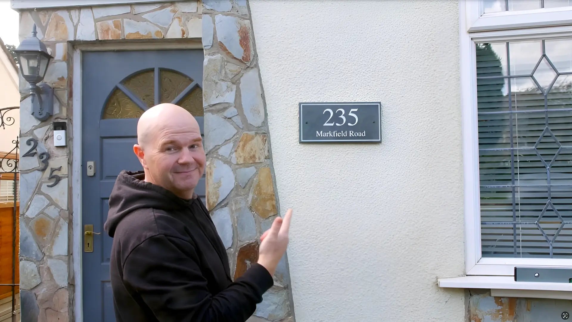 picture of a man pointing to the wall of his house. He has just installed a grey slate house sign reading 235 Markfield Road. The sign is on his beige pebble-dashed wall in between his dark grey front door and window