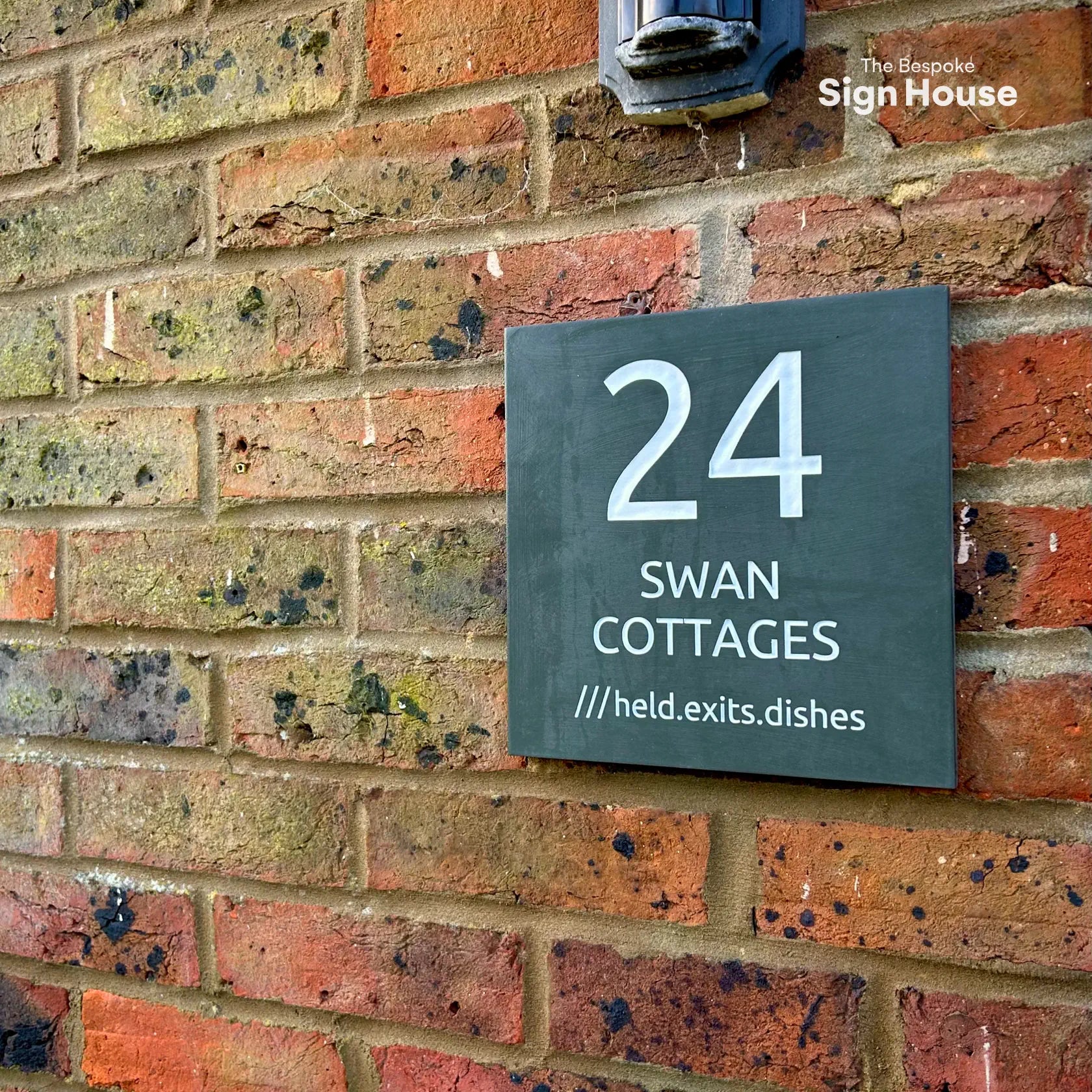 A slate house sign mounted on a brick wall, displaying “24 Swan Cottages” along with the What3Words location “///held.exits.dishes.”