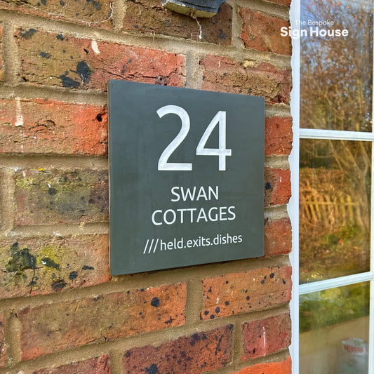 A slate house sign mounted on a brick wall, displaying “24 Swan Cottages” along with the What3Words location “///held.exits.dishes.”