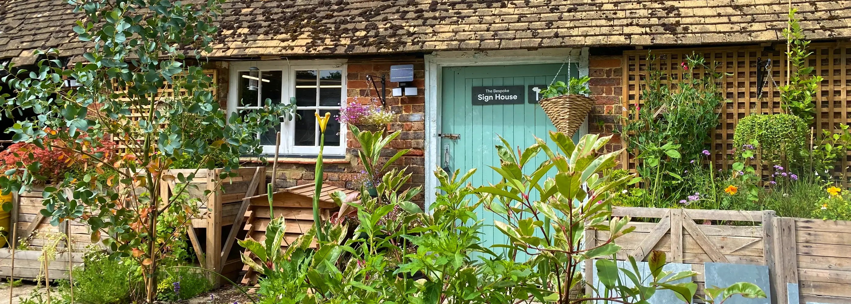 A quaint brick workshop with a sloped roof, adorned with a sign reading “The Bespoke Sign House.” The entrance has a light blue door. Surrounding the building is a lush, colorful garden with various plants and flowers, a small tree in the foreground, and wooden structures, creating a charming and vibrant atmosphere.